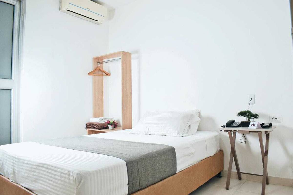 A bedroom with a bed, a bedside table with a lamp, a hanging light fixture, and a small desk with a chair.