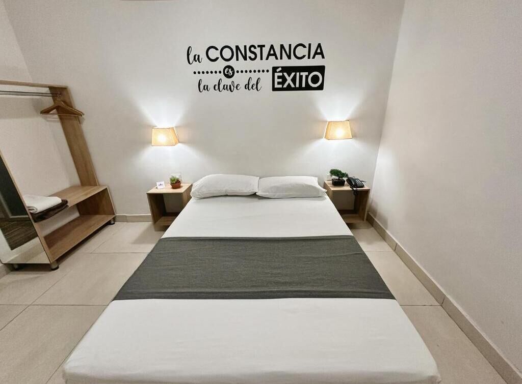 A modern hotel room with a bed, bedside lamps, a small table with a plant, and a wall-mounted sign.