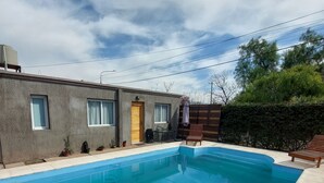 Una piscina rectangular con solárium empinado, un pequeño patio con mesa y sillas, y un edificio con fachada gris y puerta de madera.
