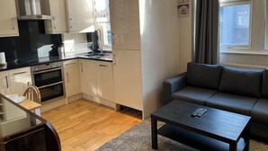 A modern kitchen with white cabinets, a black countertop, and a built-in oven. There is a grey sofa and a small table in the living area.