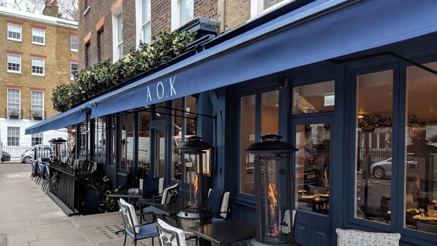 An outdoor seating area with tables and chairs, a blue awning with the letters "A.O.K.", and a fire feature in the center.