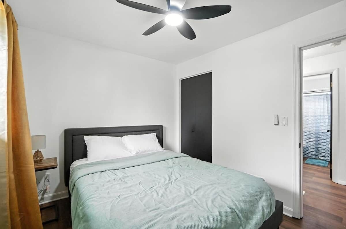 A bedroom with a bed, a ceiling fan, a nightstand, and a door leading to another room.