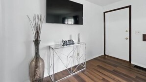 Ein Flur mit Holzboden, einer weißen Wand, einem schwarzen Fernseher, der über einem Glastisch montiert ist, und einer Tür mit dunklem Rahmen.