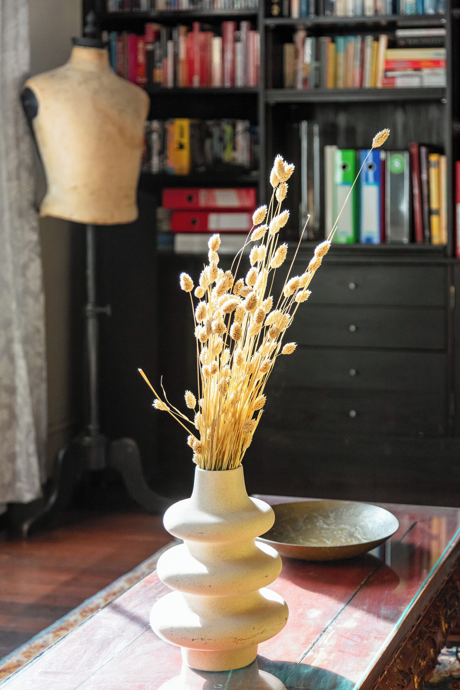 En vase med visne blomster på et bord, en lampe og en reol i baggrunden.