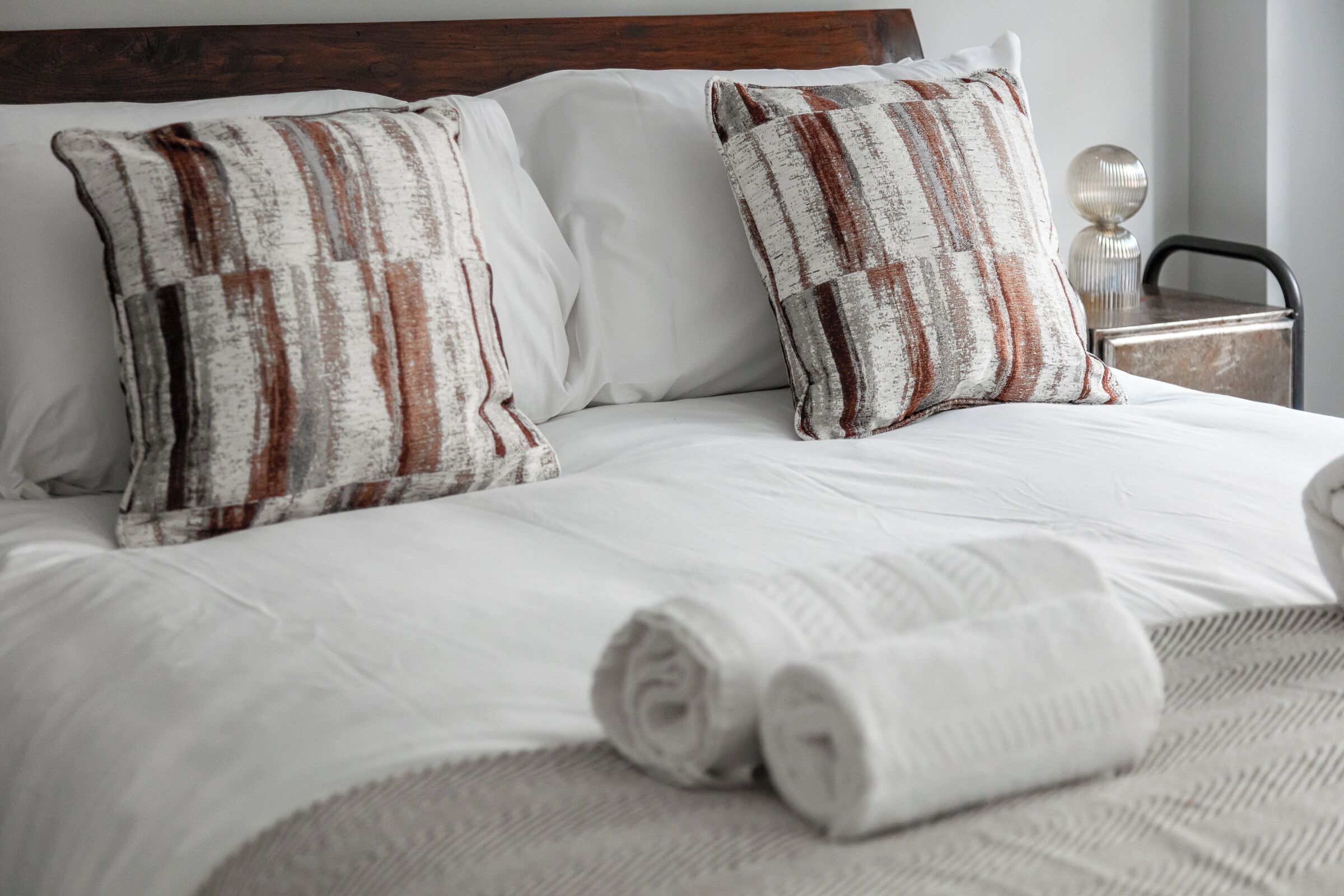 Un lit bien fait, avec du linge de lit blanc et des oreillers à rayures, une serviette blanche roulée et une table de chevet avec une lampe.