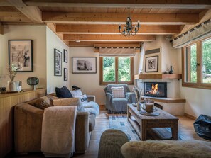 Una sala de estar acogedora con chimenea, vigas de madera y un ventanal con vistas a árboles.