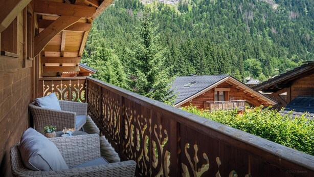 Ein hölzerner Balkon mit Korbmöbeln, der auf eine gebirgige Landschaft mit dichten Wäldern blickt.