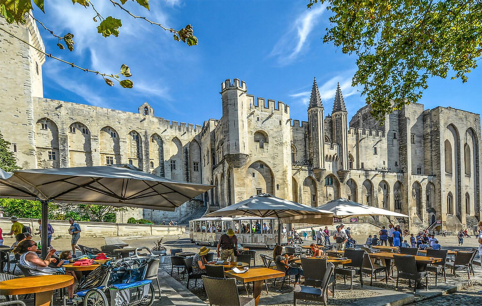 Un café al aire libre con mesas y sombrillas frente a un edificio histórico con ventanas arqueadas y torres.