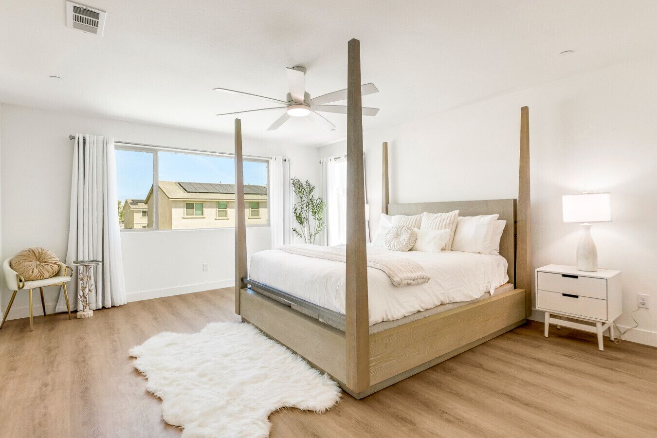 Un lit à baldaquin avec une literie blanche, une table de chevet blanche, une lampe blanche et un tapis blanc et moelleux dans une chambre dotée d’un plancher en bois et d’une grande fenêtre.