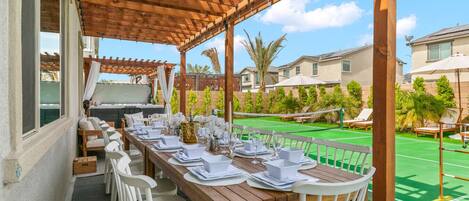 Un área de comedor al aire libre con una pérgola de madera, una mesa de billar y una cancha de tenis al fondo.