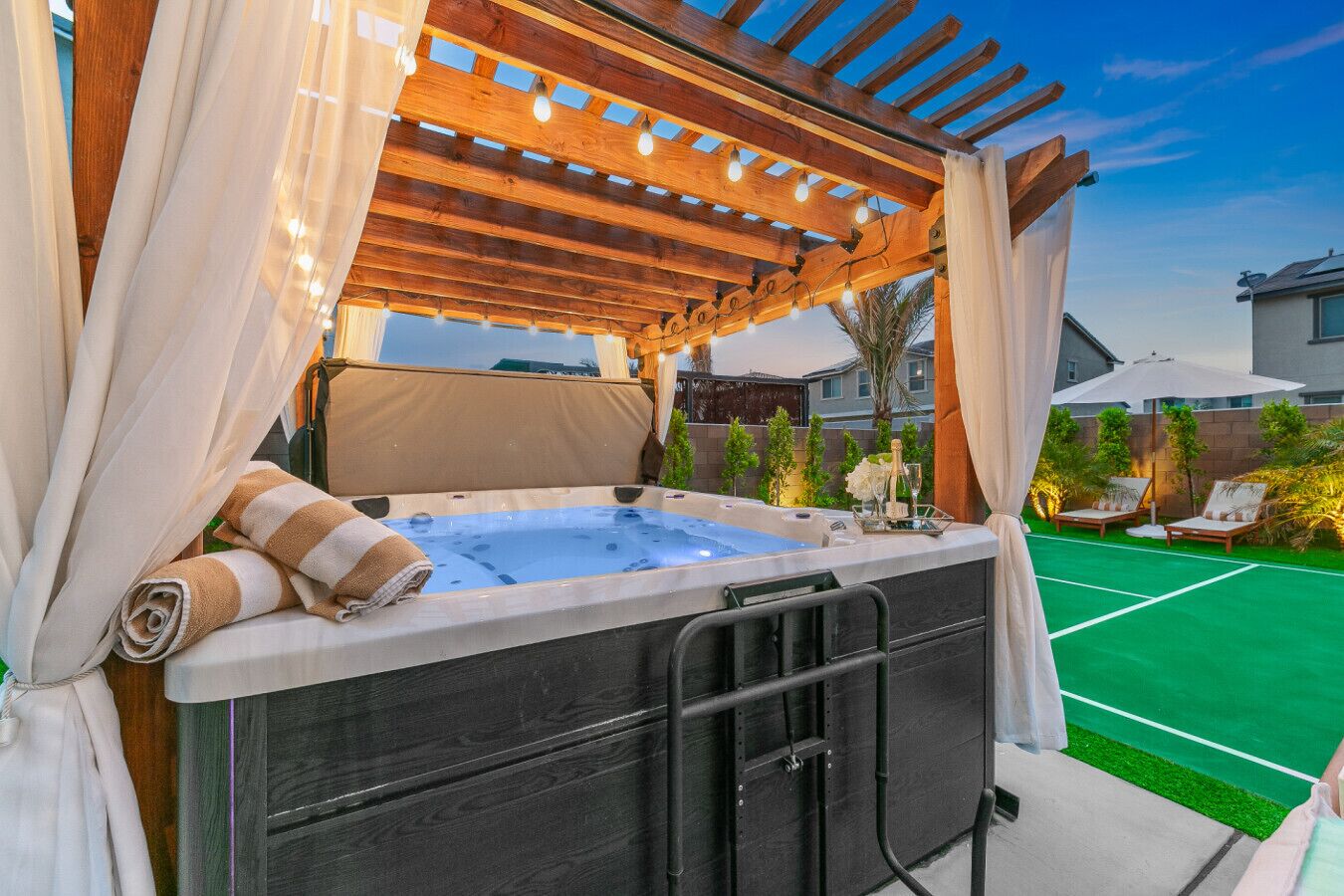 A hot tub under a wooden pergola with a view of a tennis court and a house in the background.