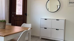 Un coin repas avec une table en bois et des chaises blanches, un meuble blanc avec des tiroirs, un miroir rond accroché au mur et une porte dotée d’un panneau en verre dépoli.