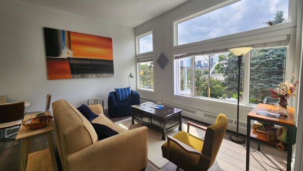 Ein modernes Wohnzimmer mit Sofa, Sessel und Couchtisch. Ein großes Fenster bietet Blick auf Bäume und Gebäude.