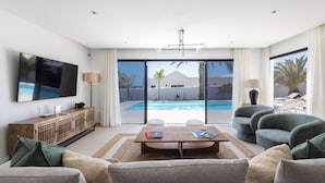 Un salón moderno con televisor de pantalla plana, una mesa de centro de madera y vista a la piscina a través de amplias puertas correderas.