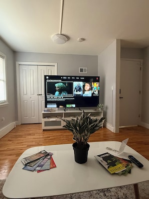 A living room with a flat-screen TV, a coffee table with magazines and a plant, and a door in the background.