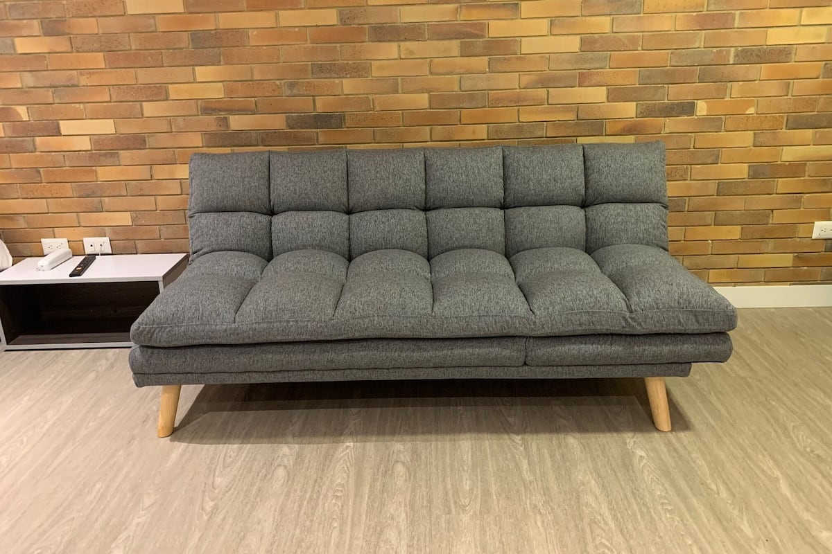 A grey sofa with wooden legs in front of a brick wall.