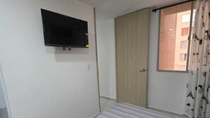 A bedroom with a bed, a TV mounted on the wall, a window with curtains, and a door leading to another room.