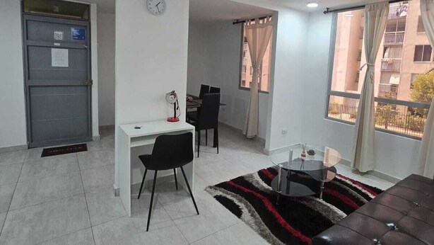 A modern apartment with a dining area, a living room with a sofa, a small desk, and a window with curtains.