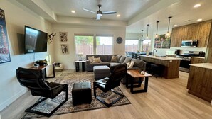 A spacious living room with a large sofa, a coffee table, and a TV mounted on the wall. There is a kitchen area with a microwave, stove, and bar stools.