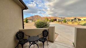 A patio with two ornate metal chairs and a table, a beige wall, and a view of a desert landscape with mountains in the distance.