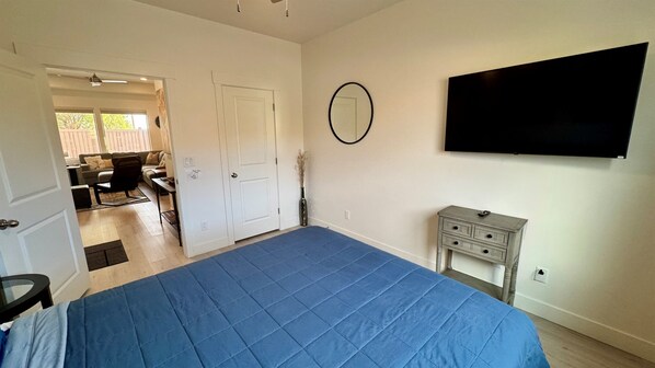 A bedroom with a large bed, a TV mounted on the wall, a small bedside table, and a door leading to a living area. - Desert Basecamp near Zion & Gooseberry Mesa | Family-Friendly New Build (Apple Valley)