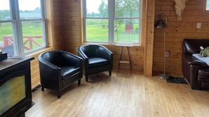 L’intérieur d’un chalet en bois, avec deux fauteuils en cuir, une table en bois, une cheminée et une grande fenêtre donnant sur un paysage verdoyant.