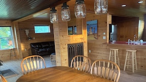 L’intérieur d’un chalet en bois comprend une table à manger, des chaises, une cheminée, un coin cuisine et un salon avec un canapé en cuir noir.