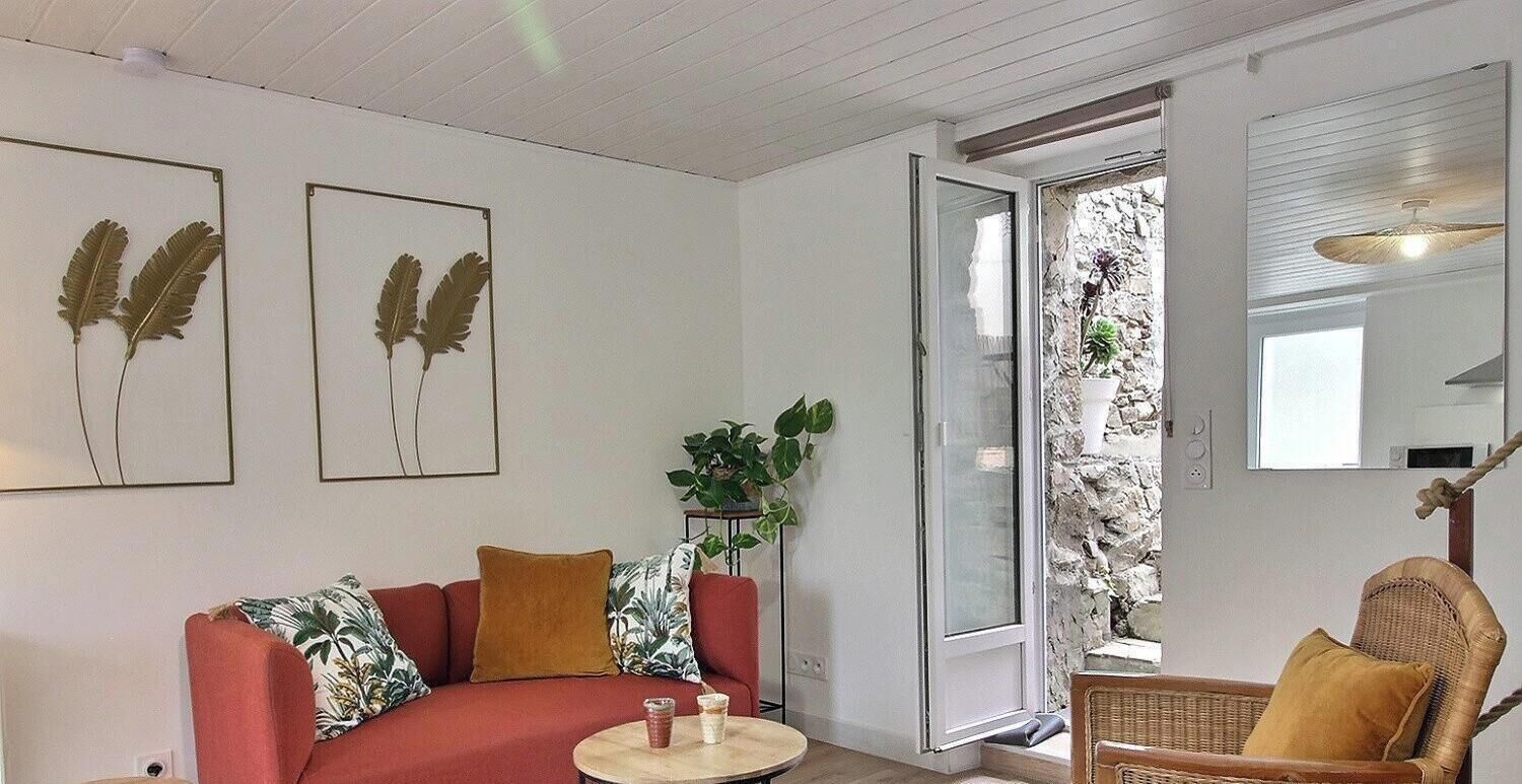 Ein Wohnzimmer mit einem roten Sofa, einem Korbstuhl, einem kleinen runden Tisch und einer Obstschale. An der Wand befinden sich zwei gerahmte Blätter-Dekorationen.
