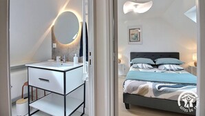Un dormitorio con cama, mesitas de noche y un baño con lavabo y espejo.