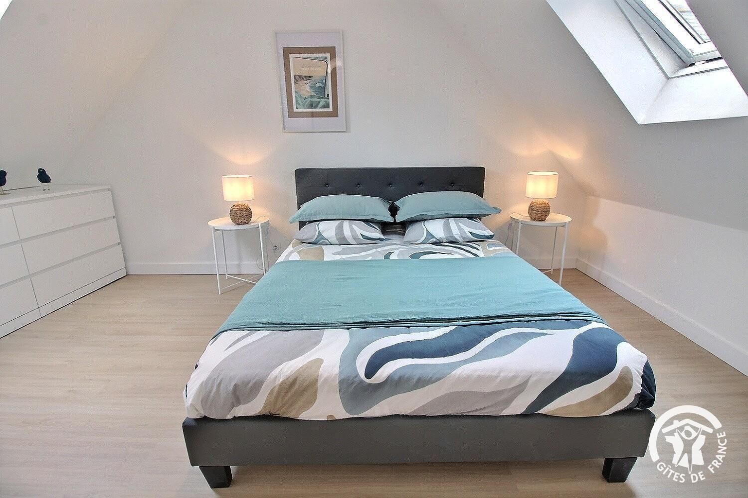 A bedroom with a bed, two bedside tables, a nightstand, a dresser, a framed picture on the wall, and a skylight.