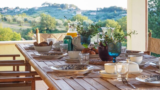 Une table de salle à manger en bois, mise avec des assiettes, des verres et un vase rempli de fleurs, donnant sur un paysage pittoresque.