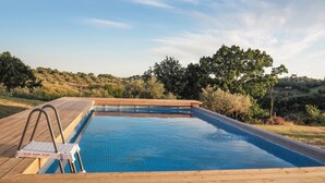 Une piscine rectangulaire dotée d’une terrasse en bois et d’un escalier en métal, entourée de verdure et de collines.