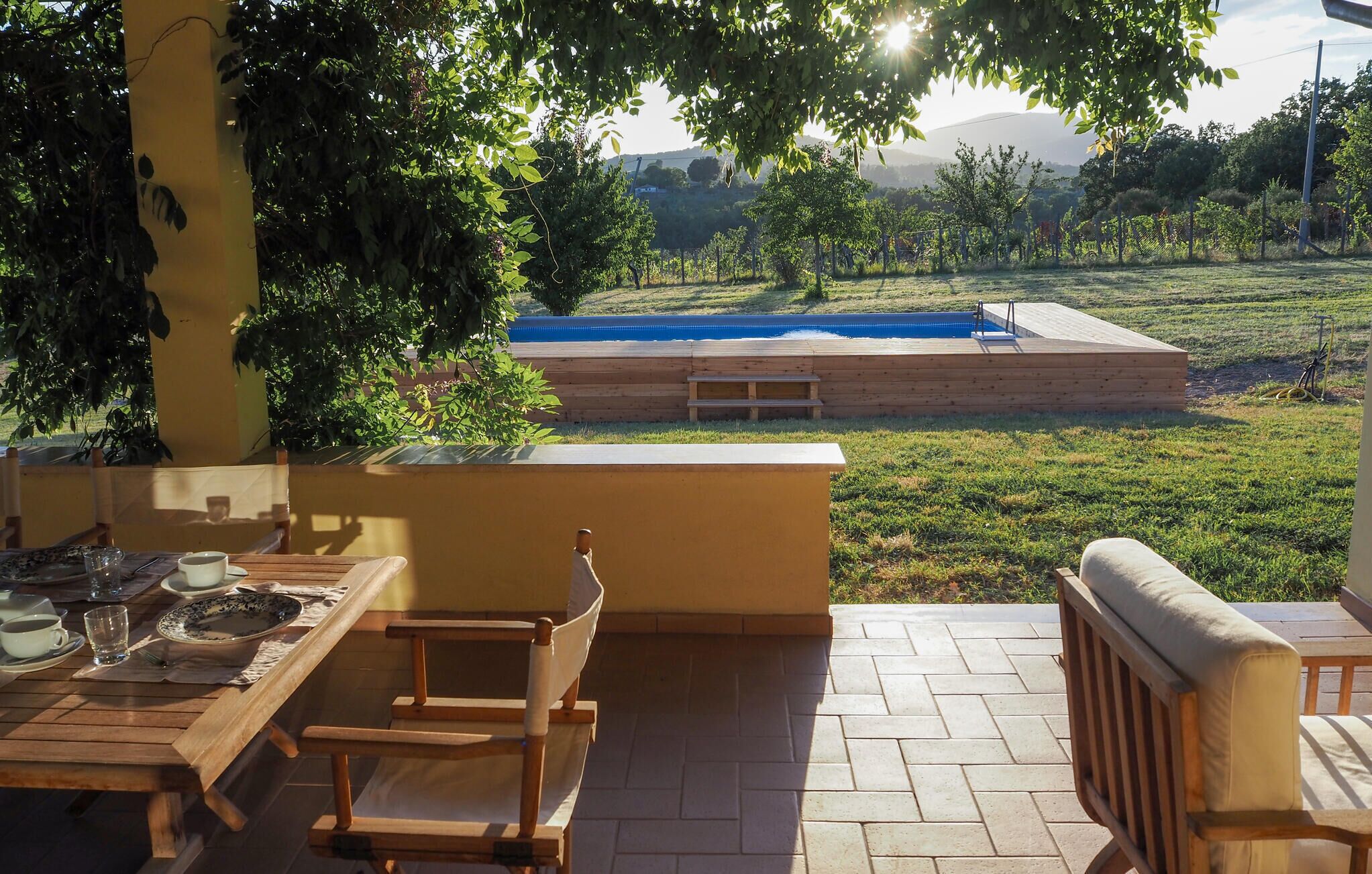 An outdoor patio with a wooden table and chairs, a small pool, and a view of a hilly landscape.