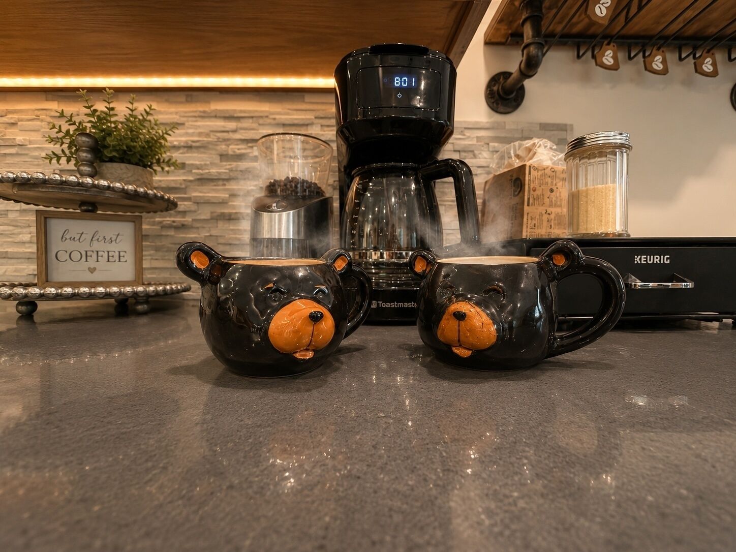 Eine Kaffeemaschine, zwei Tassen in Bärenform, ein Toaster und ein Schild mit der Aufschrift 'aber zuerst KAFFEE' auf einer Küchenarbeitsplatte.