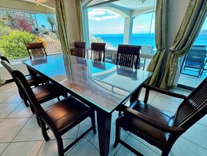 A glass-top dining table with six chairs, set in a sunlit room with a view of the ocean.