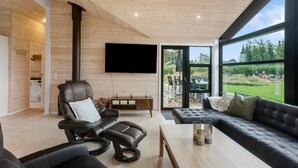 Una sala de estar moderna con un sofá de cuero, un sillón reclinable y una mesa de centro. Un televisor de pantalla plana está montado en la pared, y hay un ventanal que ofrece vistas al exterior.