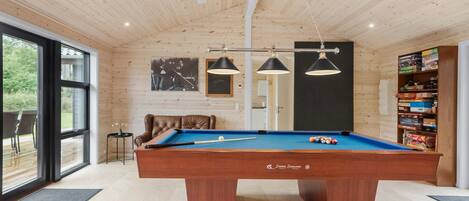 A pool table with a billiard cue, a leather chair, and a bookshelf with board games.