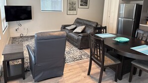 Un salon moderne avec un canapé en cuir, une table à manger avec des chaises, un téléviseur à écran plat fixé au mur et un réfrigérateur dans la cuisine.
