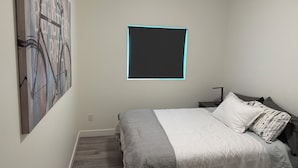 Une chambre à coucher comprenant un lit, une table de chevet, un téléviseur fixé au mur et un grand tableau abstrait.