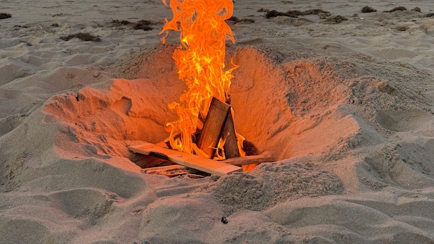 Escena de playa con un hogar a la intemperie en la arena y una persona sentada con los pies levantados.