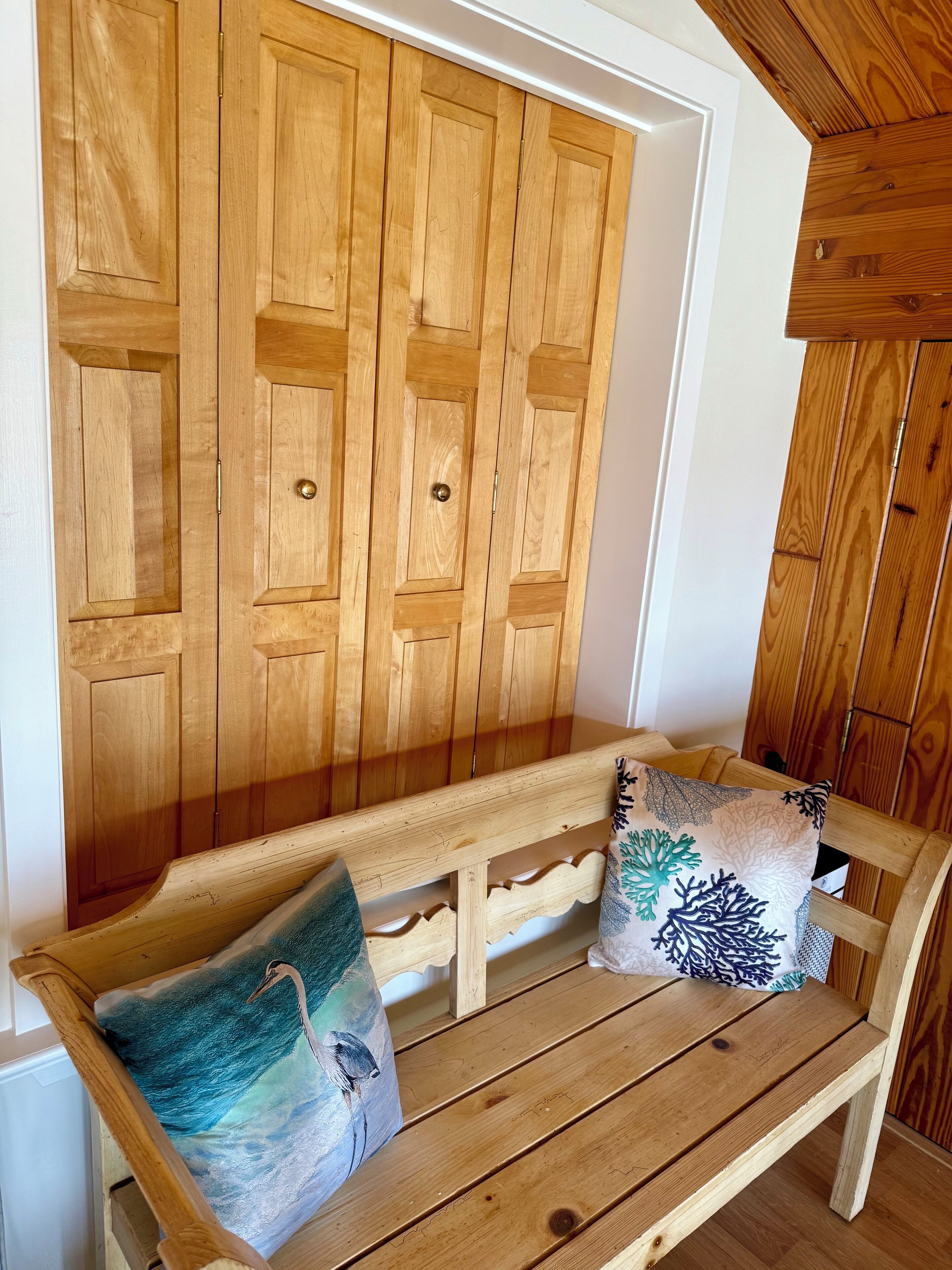 A wooden bench with two cushions, one with a bird design, and a wooden wardrobe with a mirrored door.