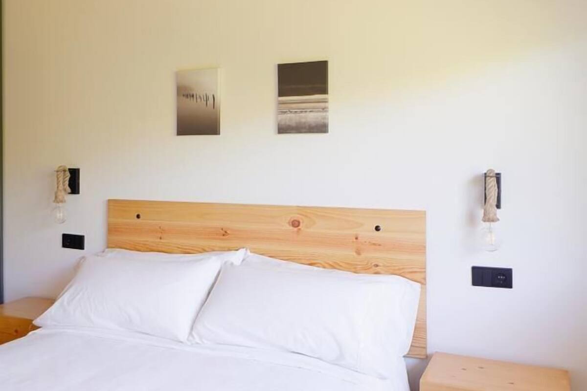 Cama com cabeceira de madeira, duas mesas de cabeceira e duas luminárias fixadas na parede.