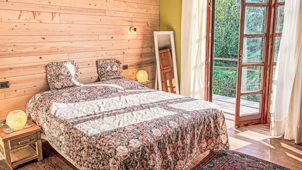 A bedroom with a wooden slat wall, a bed with floral bedding, a nightstand with a lamp, and a view of greenery through a glass door. - Del Respiro by Interhome (Asti)