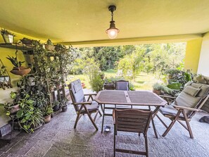 Un patio al aire libre con una mesa de madera redonda y cuatro sillas, rodeado de macetas con plantas y una pared amarilla.
