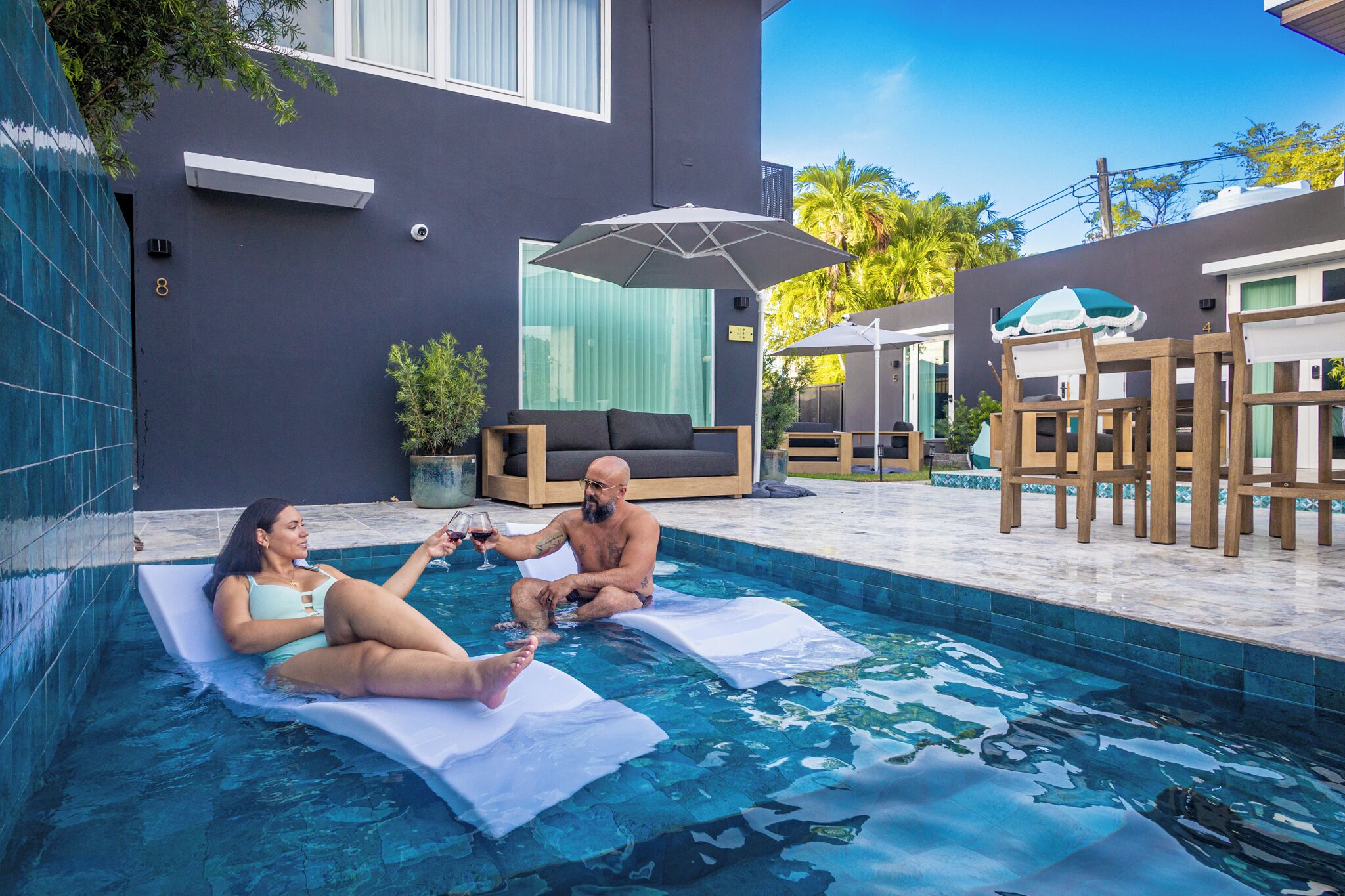 Una piscina con sillones de descanso y sombrillas, un edificio moderno con amplios ventanales y una zona ajardinada bien cuidada.