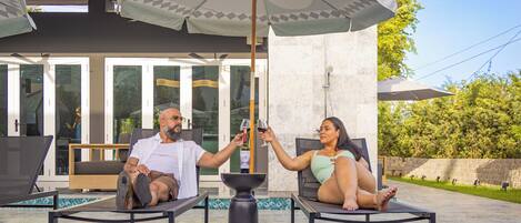 Two people relaxing on lounge chairs under a large umbrella, with a pool and outdoor seating area visible in the background.