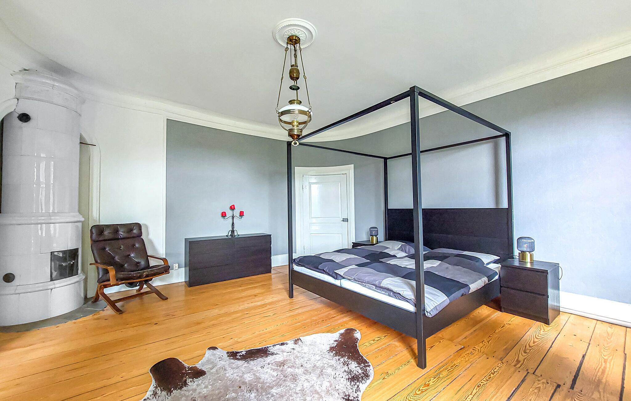 A four-poster bed with a dark headboard, a leather chair, a small table with a red candle holder, and a cowhide rug on a wooden floor.