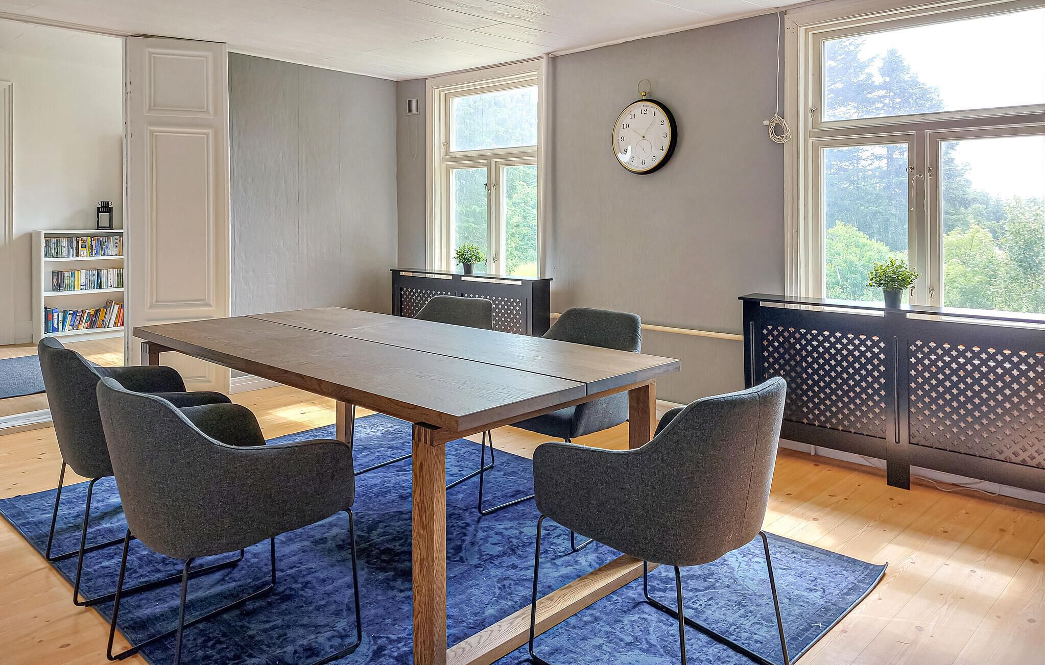 A modern dining area with a wooden table and four grey chairs, a bookshelf, and a clock on the wall.
