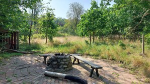 Een rustieke buitenruimte met stenen banken en een vuurkuil, omgeven door weelderige begroeiing en bomen.