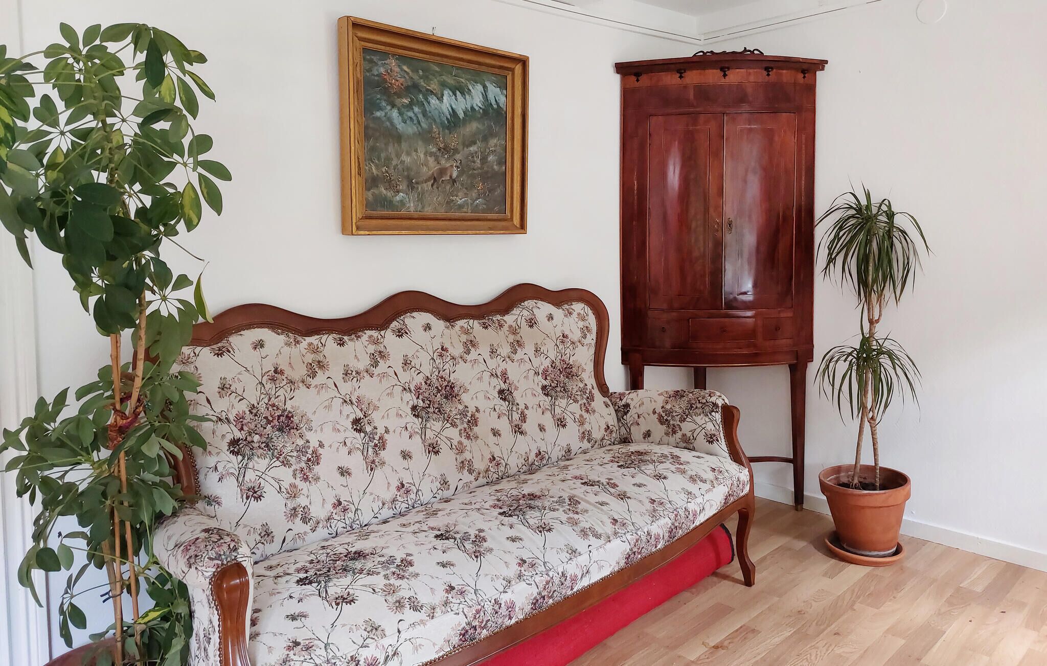A room with a floral sofa, a wooden cabinet, a potted plant, and a framed painting on the wall.